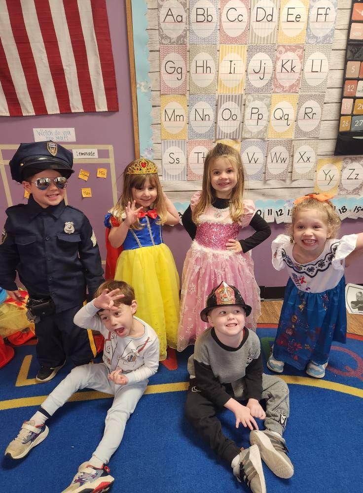 Children in costumes: policeman, Snow White, princess, superhero, firefighter and a Disney character smiling in a classroom.