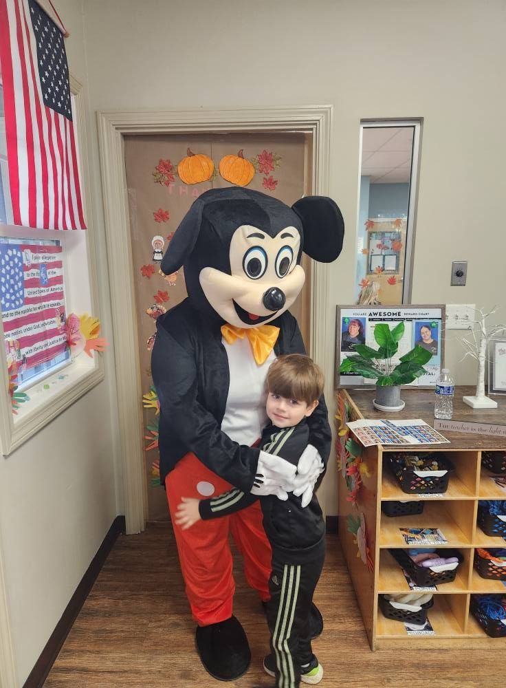 Mickey Mouse hugs a child indoors by a classroom door. American flag and fall decorations visible.