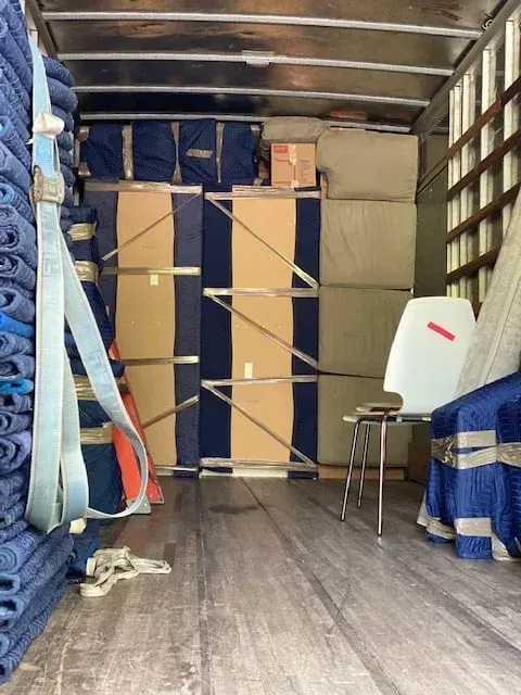 Cargo truck interior packed with stacked blue and tan boxes and rolled textiles, with straps securing the load