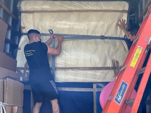 Person securing a large wrapped item in a moving truck with orange ladder and stacked boxes