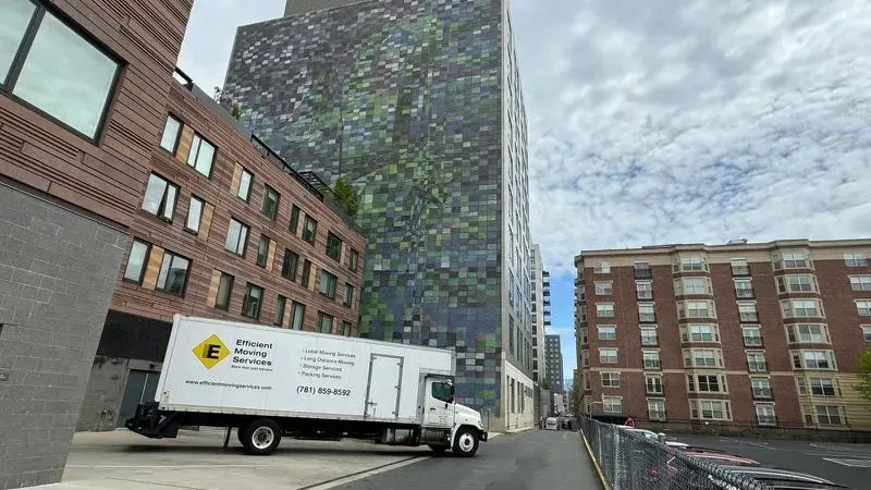 White box truck parked beside modern brick and glass buildings under a cloudy sky.