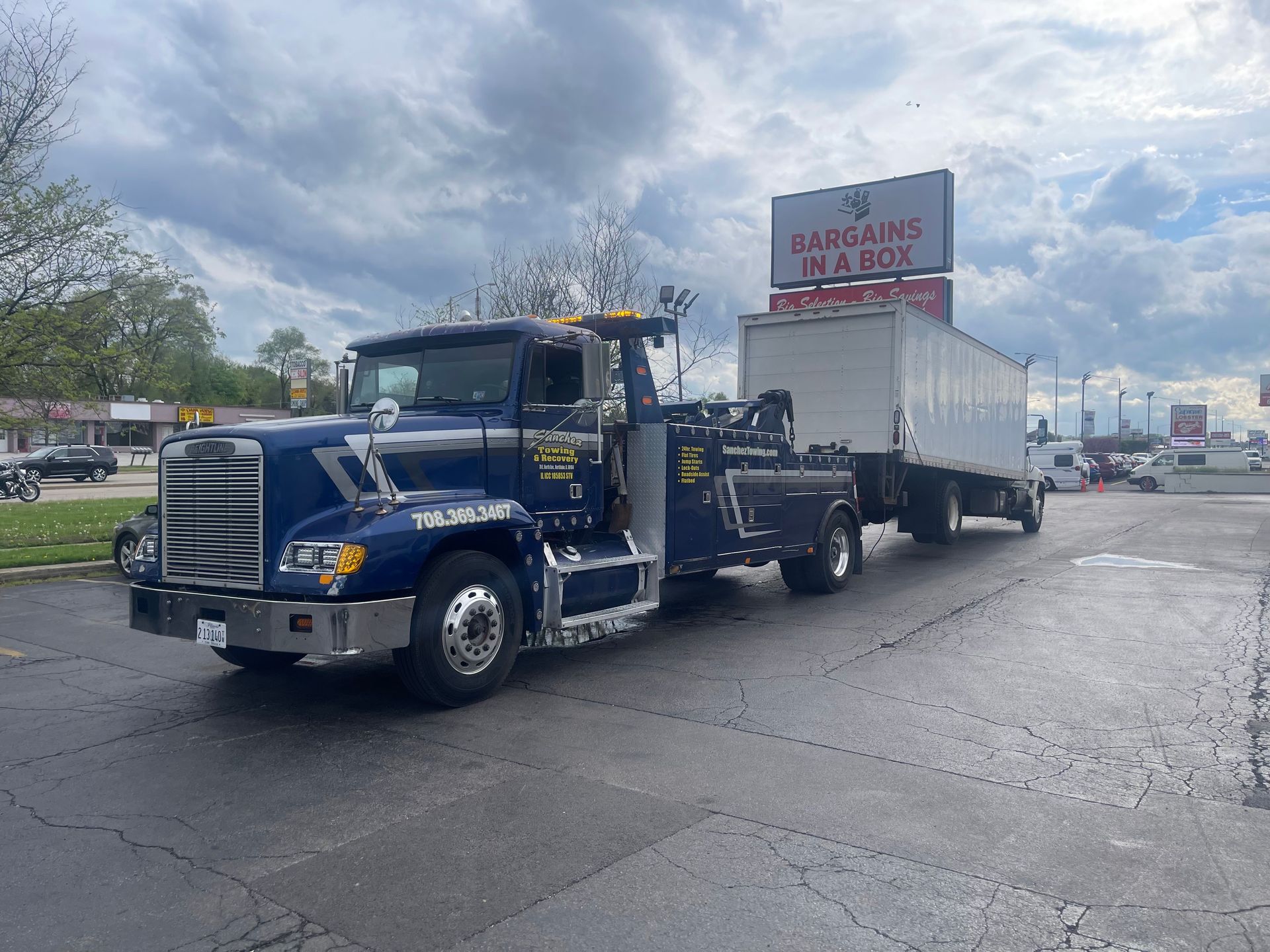 A blue tow truck is towing a white trailer in a parking lot.