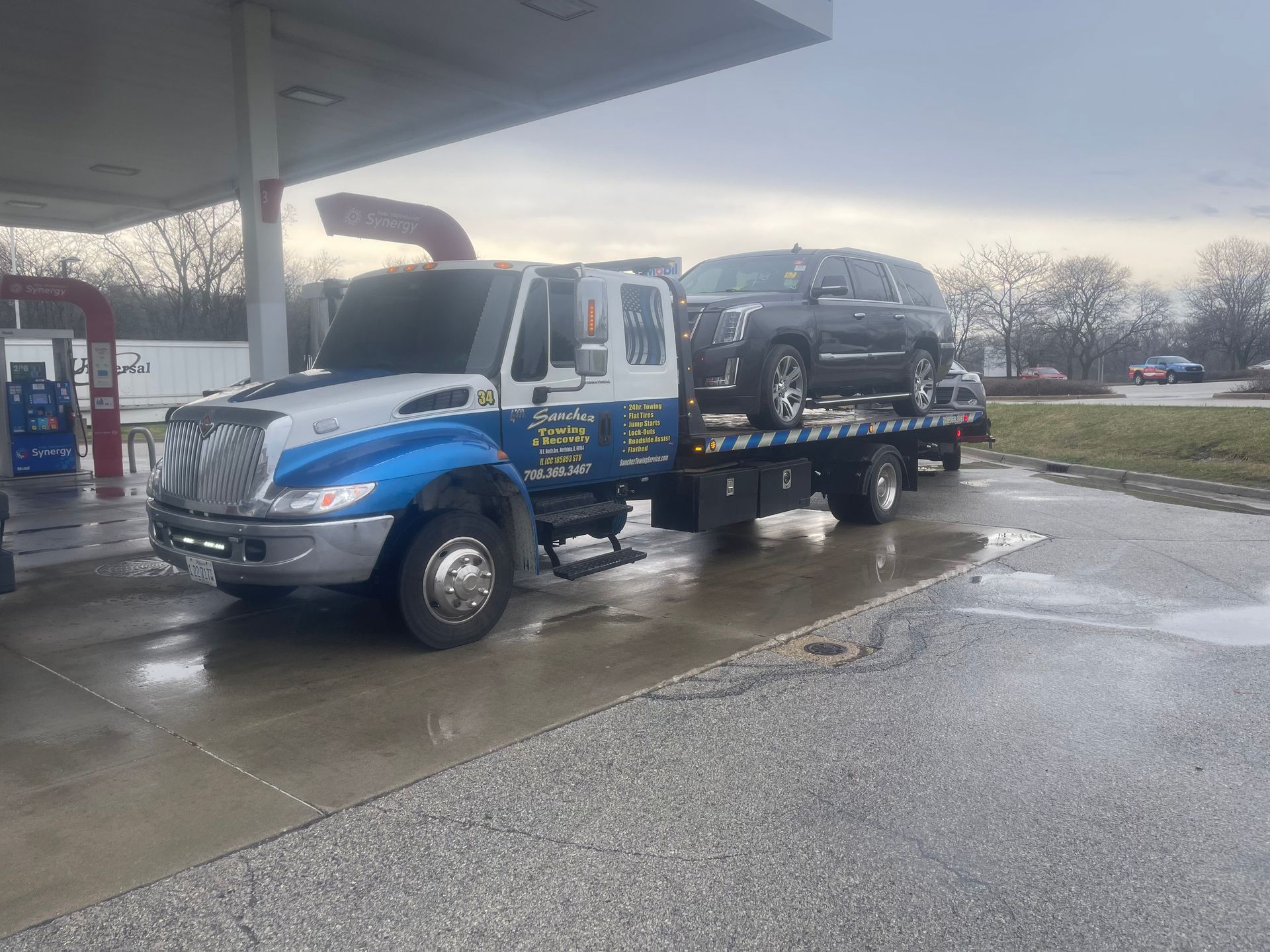 A tow truck with two cars on the back is parked in front of a gas station.