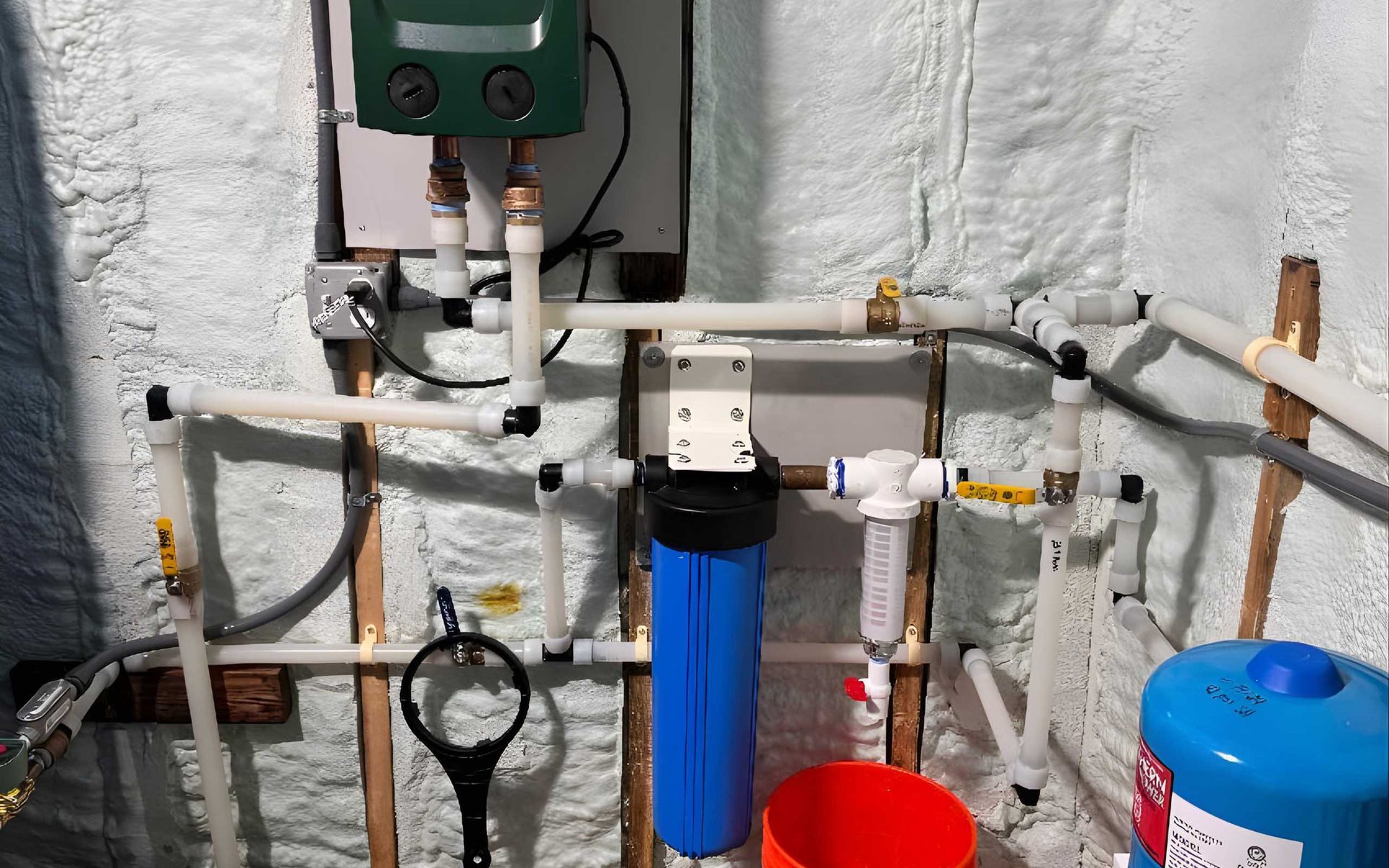 Plumbing system with filter, boiler, and pipes against insulated wall. Blue, white, copper, and black components.