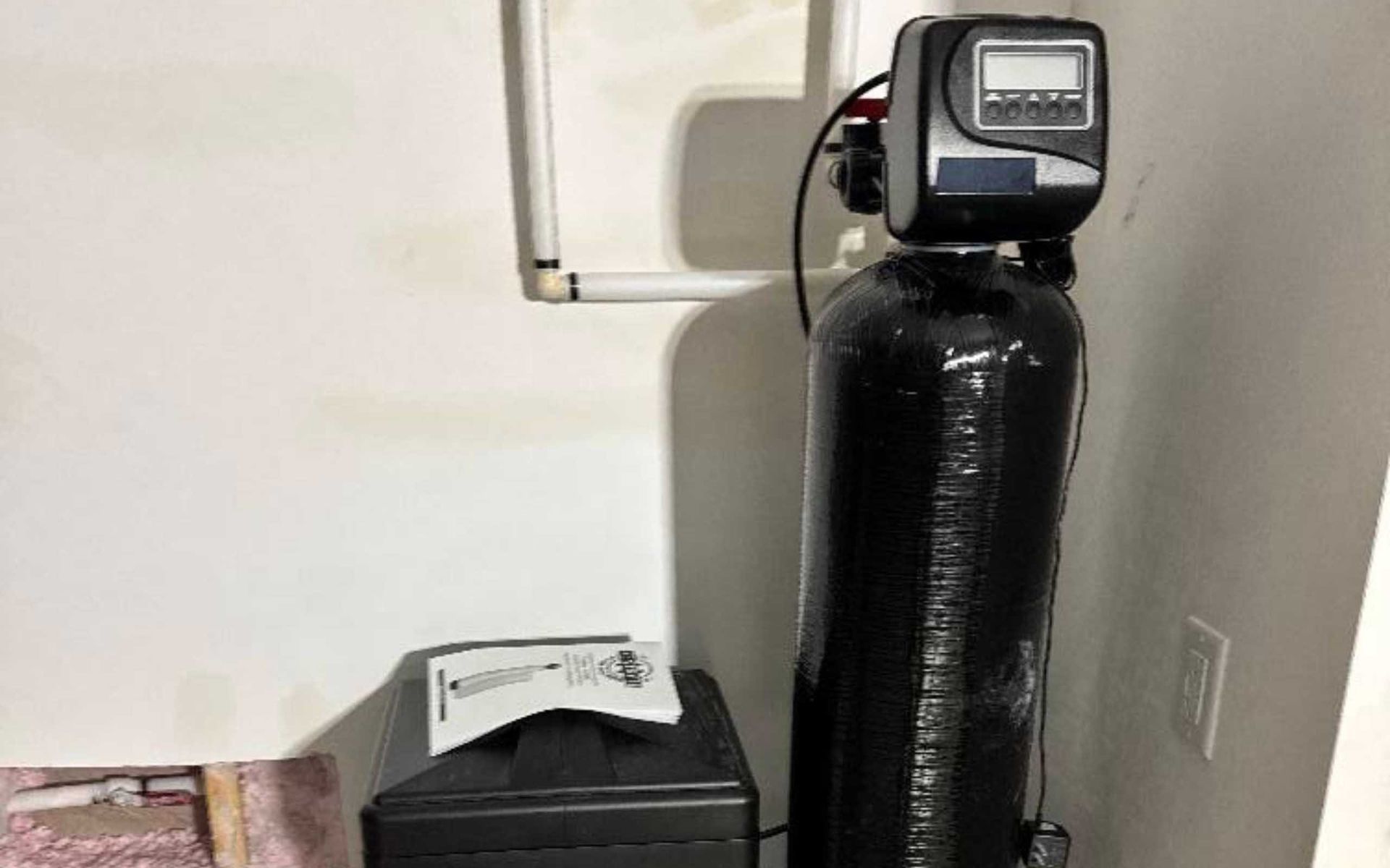 Water softener system in a utility room; black tank, digital control panel, salt tank, white walls.