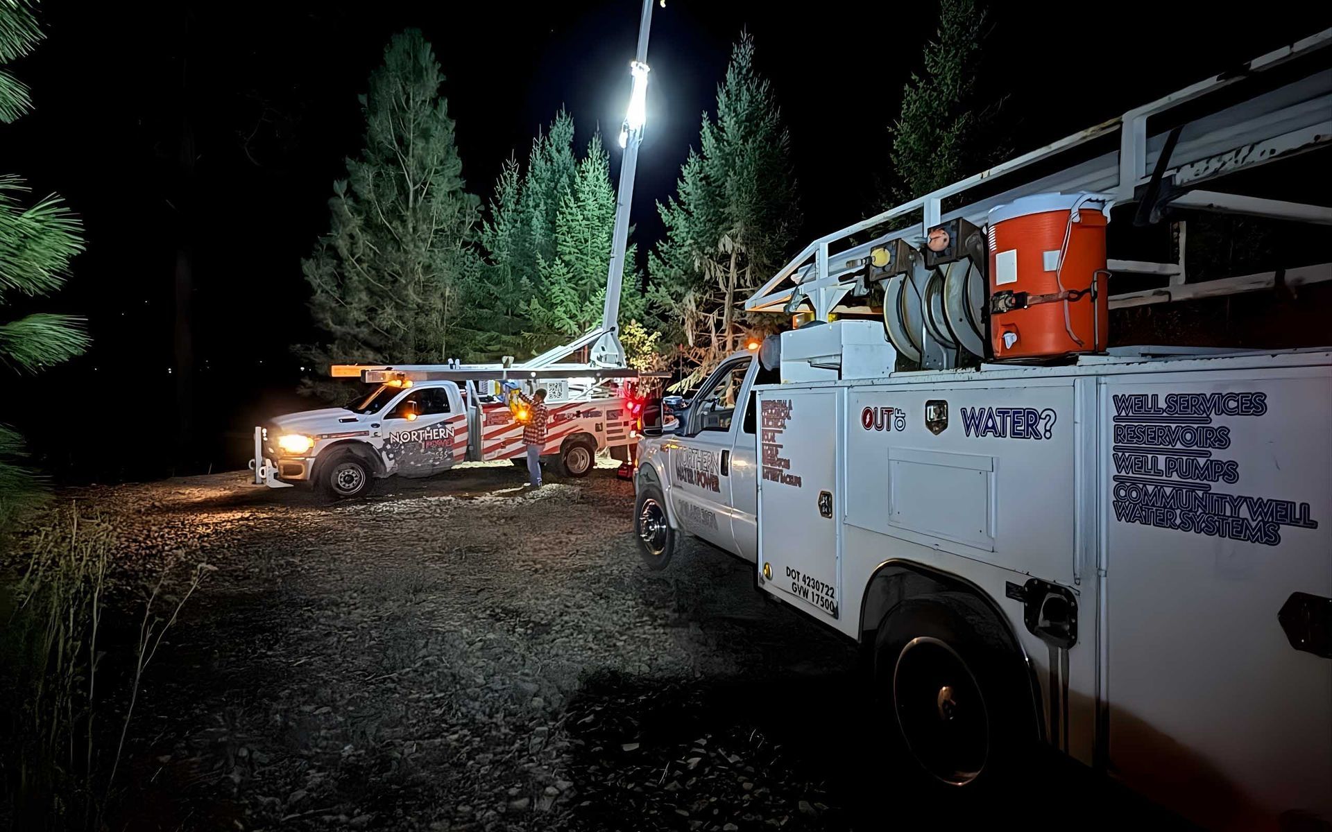 Two utility trucks with lights on at night; one has a lifted boom near trees.