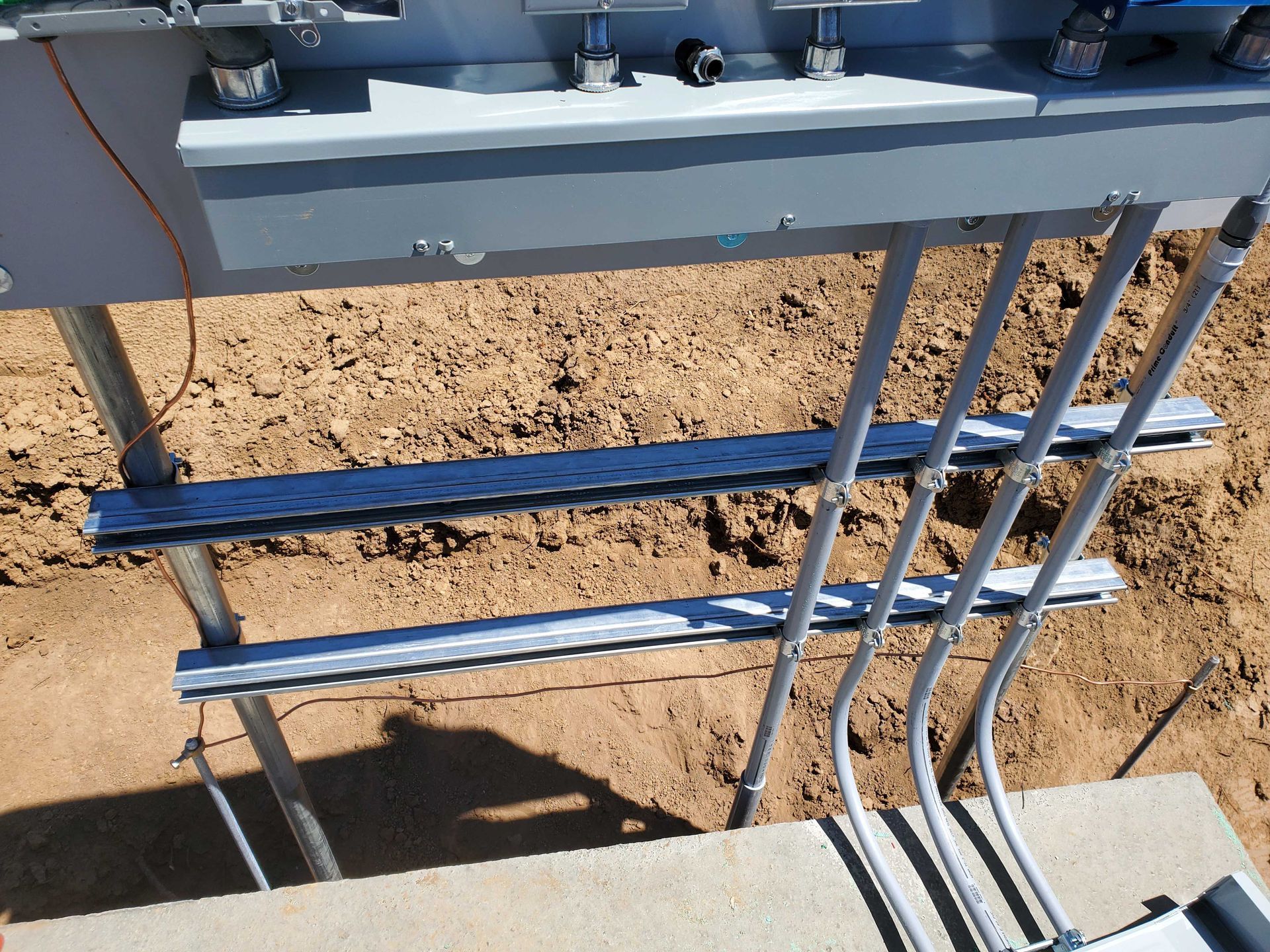 Electrical conduits run into a gray box. Metal supports and dirt are in the foreground.