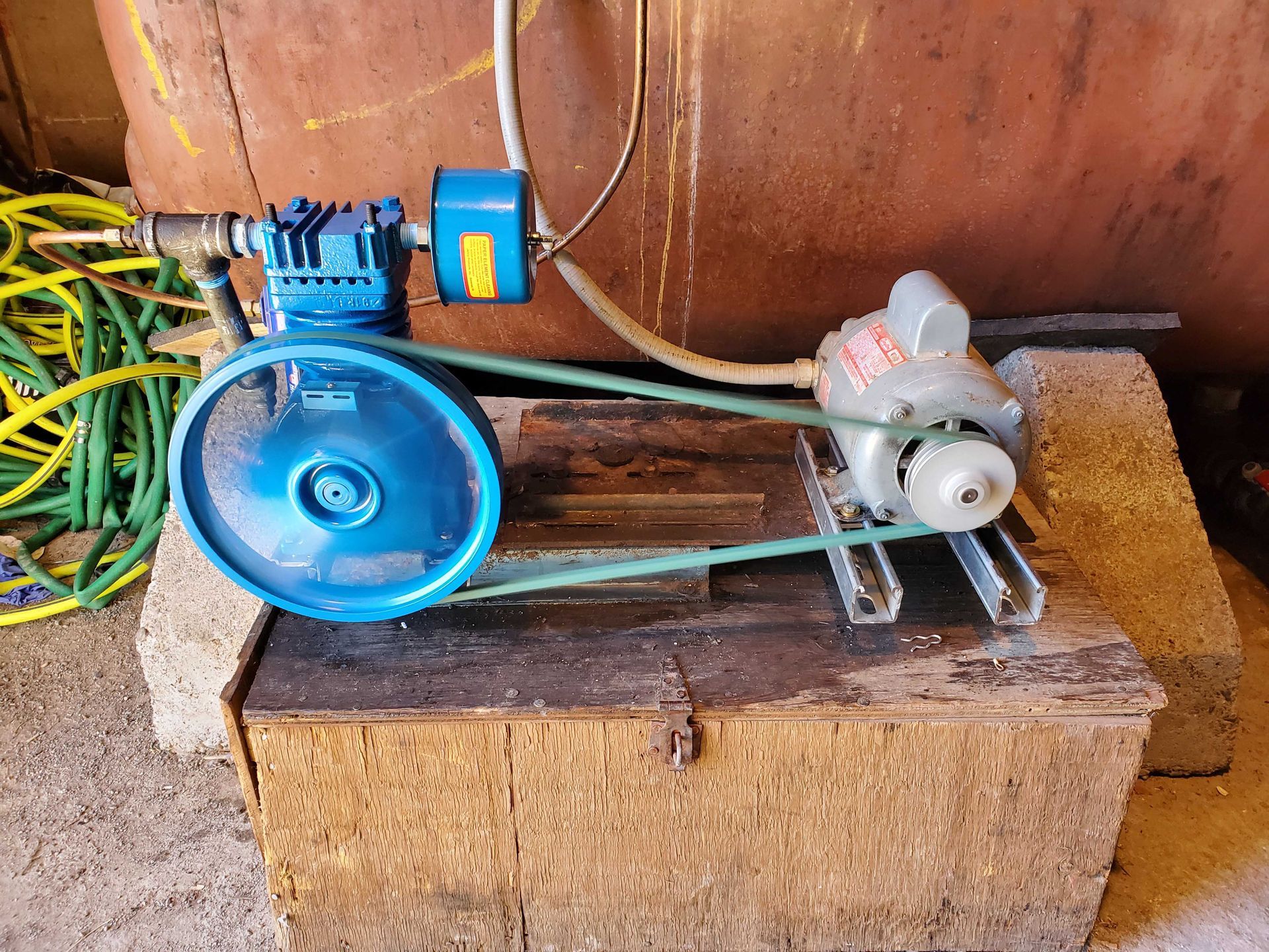 Blue air compressor with belt and motor, sitting on a wooden box.