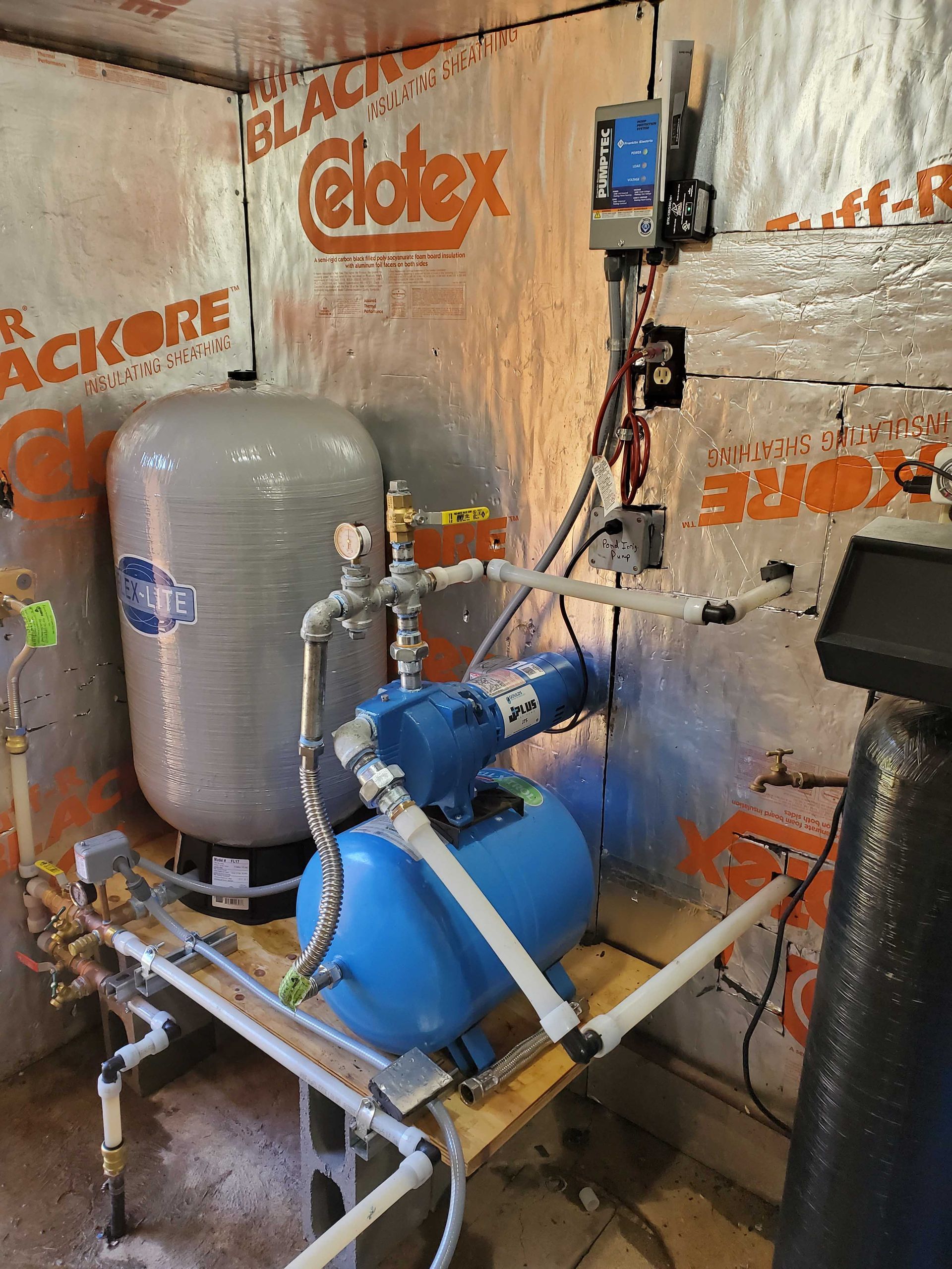 Water well pump system with pressure tank, pipes, and control panel in a small room.