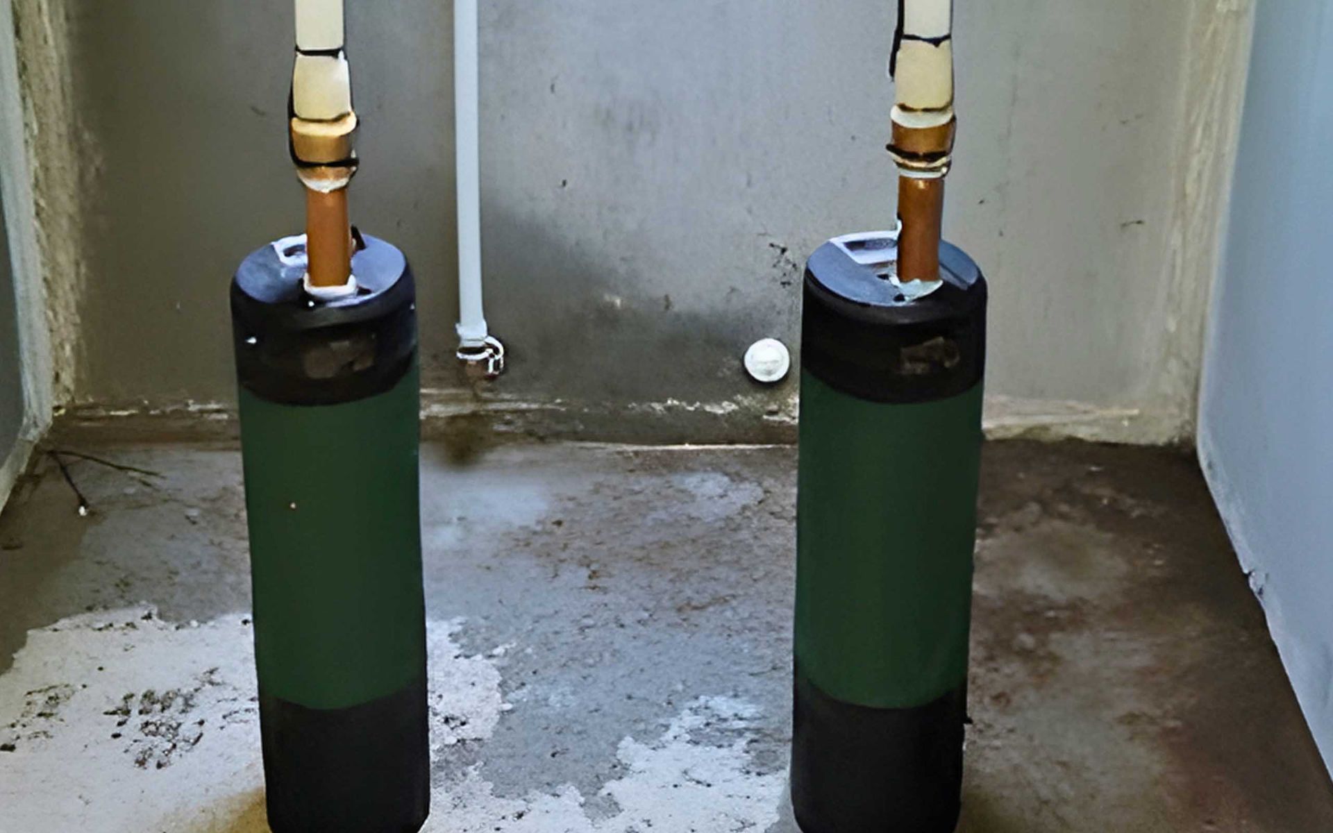 Two cylindrical water tanks, green and black, with copper pipes, stand on a concrete floor in a room.