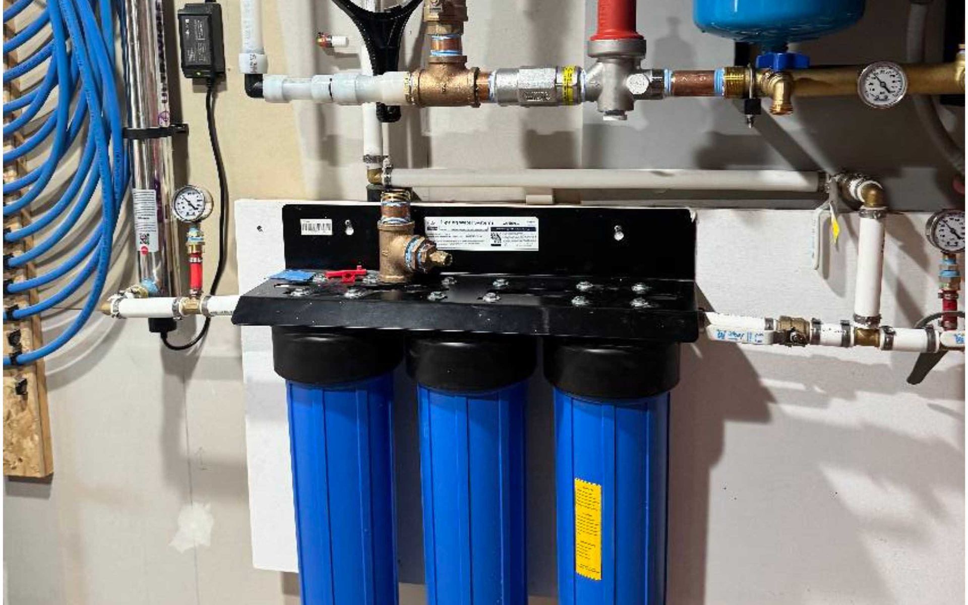 Three blue water filter housings mounted on a wall, with connected pipes and gauges.