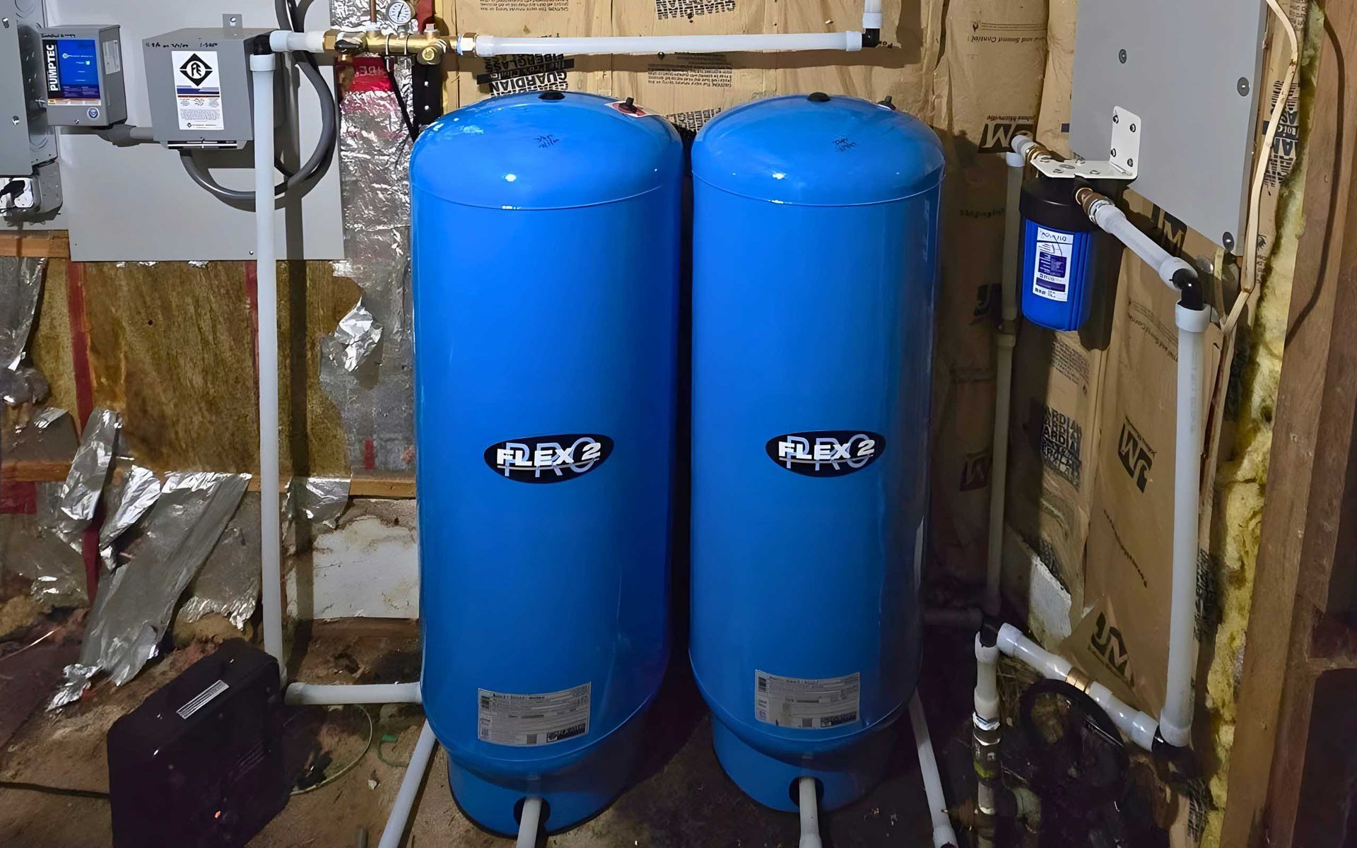 Two blue water tanks in a utility room, connected by white pipes. Wooden walls.