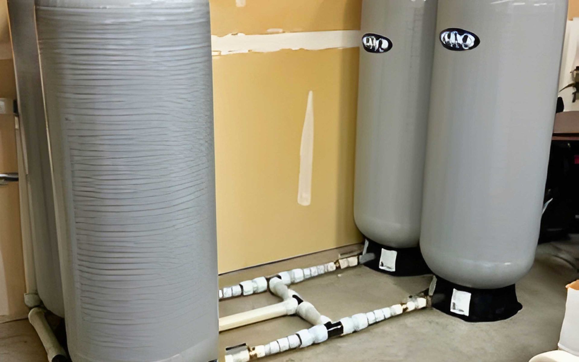 Four cylindrical water tanks with white pipes connected in a basement setting.