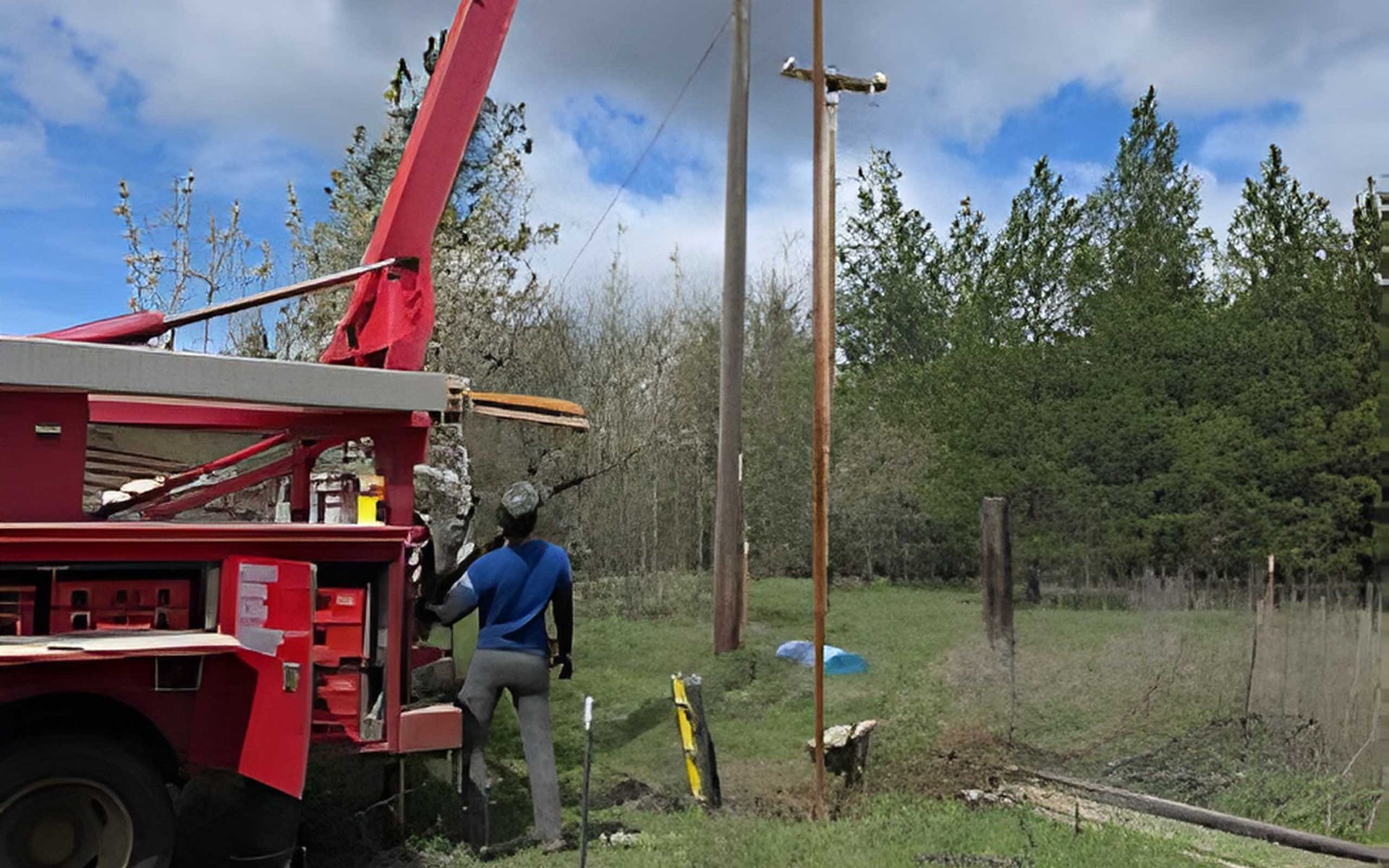 A worker by a red truck with a crane is near a utility pole in a grassy area with trees.