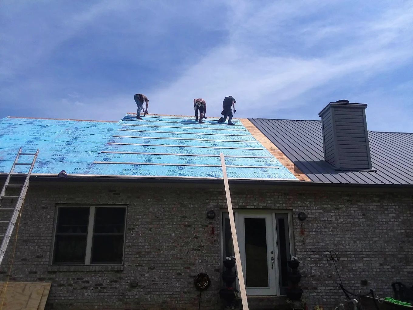 Workers installing roofing on a brick house under a blue sky.