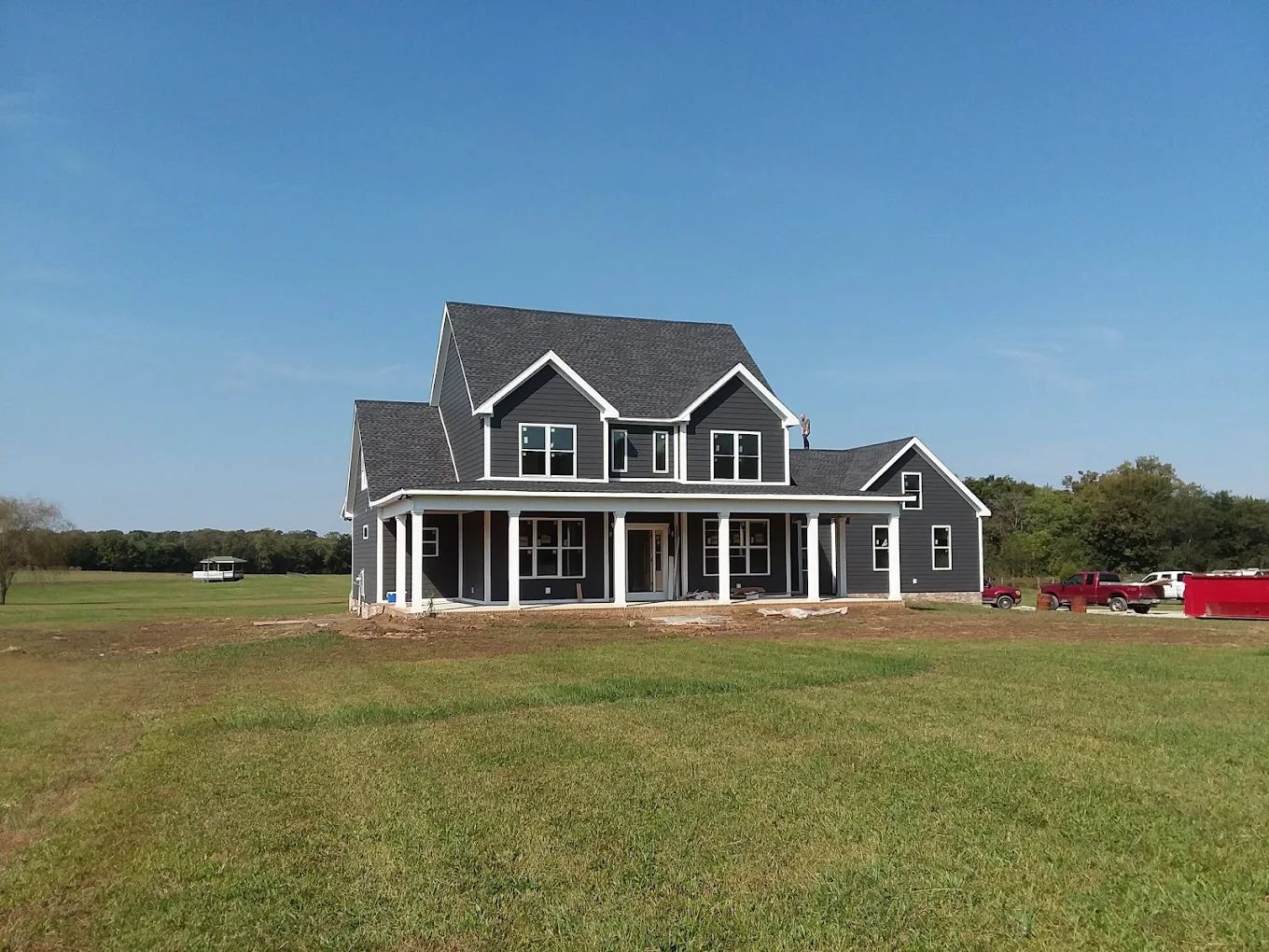 Newly constructed two-story house with dark gray siding, white trim, and a wrap-around porch in a grassy field under a blue sky.