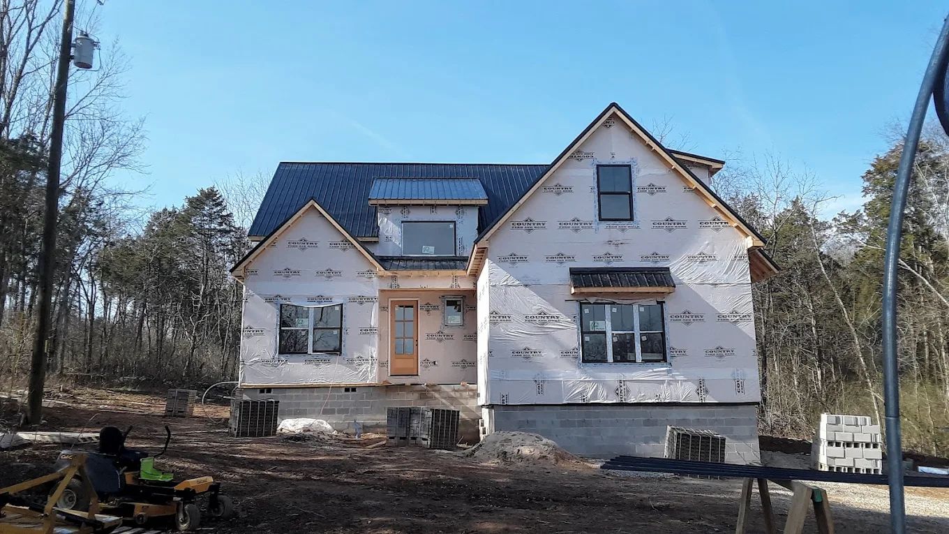 House under construction, covered in white wrap, with exposed wood framing and windows, set in a wooded area.