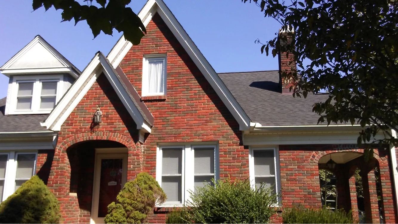 Red brick house with dark roof and white window frames. Green bushes and tree in front.