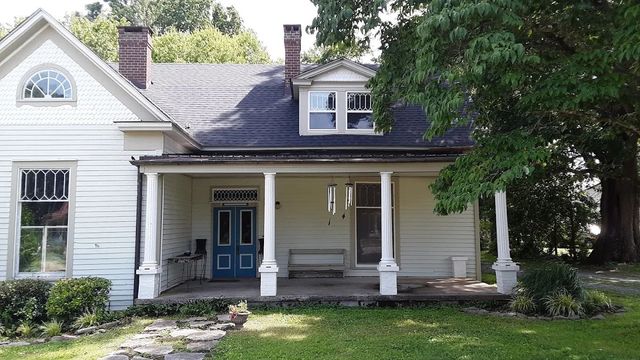 White two-story house with porch. Blue door, arched window, dark roof, trees, and green grass.