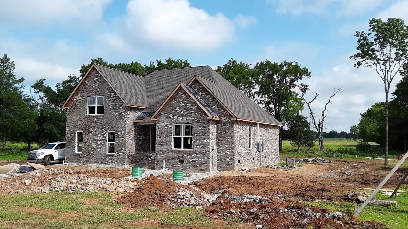 New brick house under construction, surrounded by dirt and trees, sunny day.