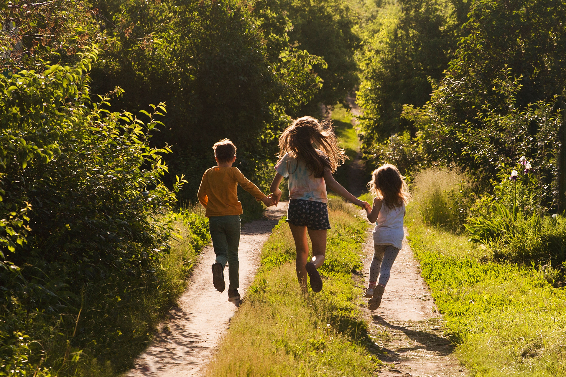 Three children are running down a dirt road holding hands.