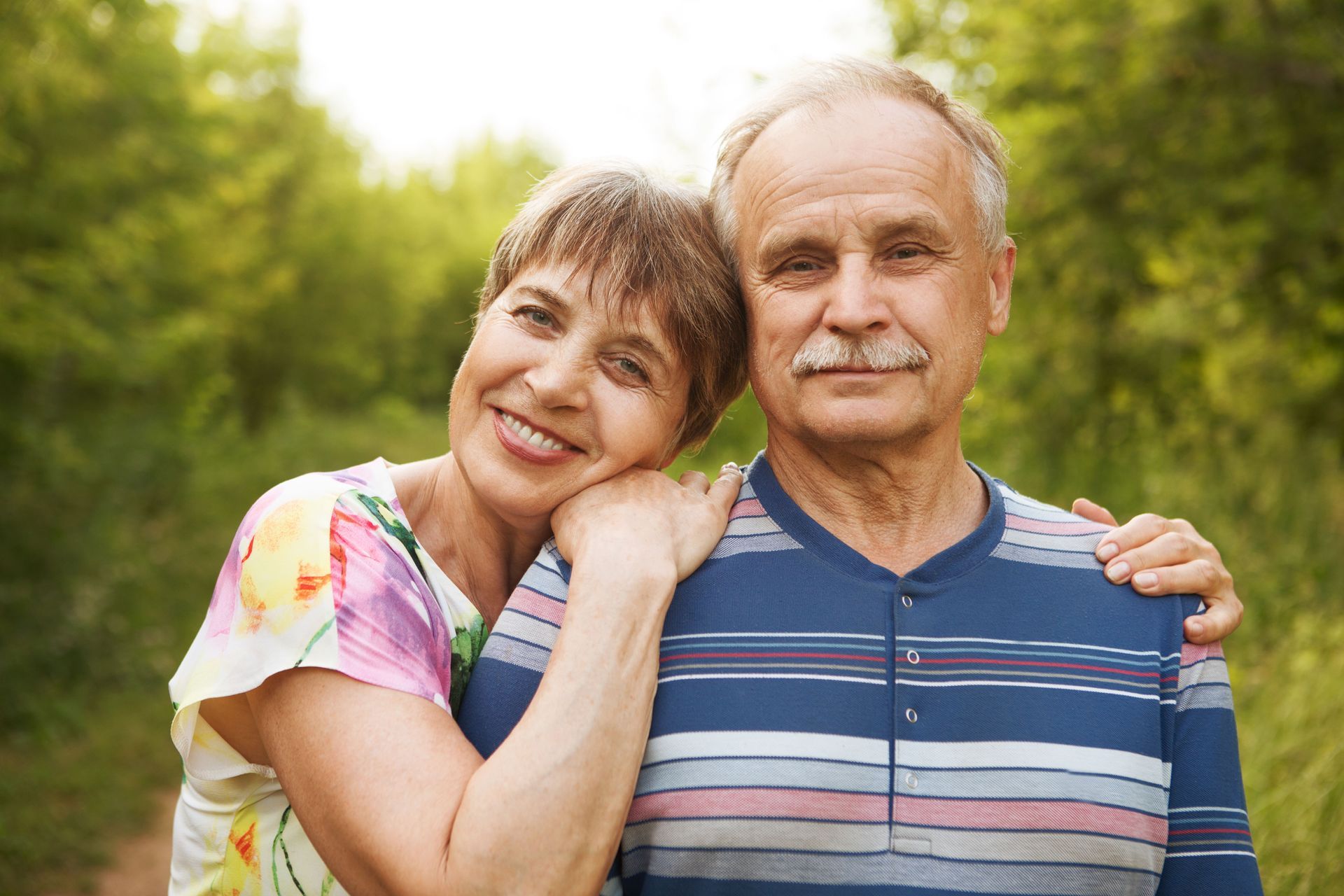 An elderly couple is posing for a picture in the woods.