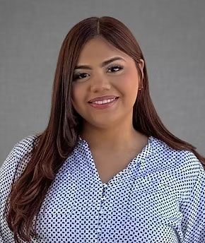 Smiling woman with long brown hair wearing a blue patterned blouse against a gray background