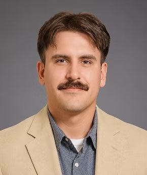 Portrait of a man with a mustache wearing a beige blazer and blue shirt against a gray background