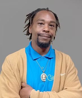 Portrait of a smiling man in a blue polo and tan cardigan against a gray background