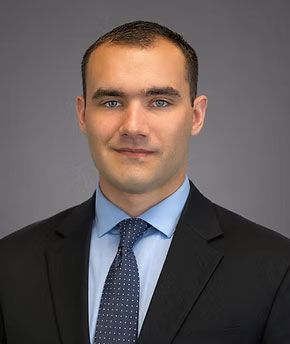 Studio headshot of a man in a dark suit and blue shirt against a gray background