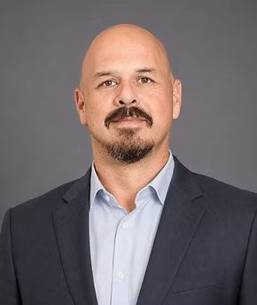 Studio headshot of a bald man with a goatee in a dark blazer and light blue shirt on a gray background