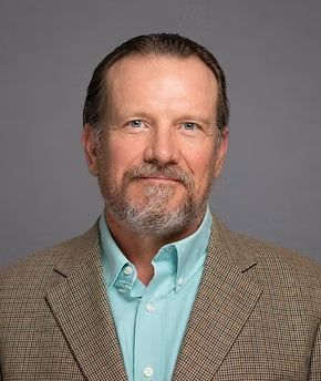Portrait of a man in a brown blazer and turquoise shirt against a gray background