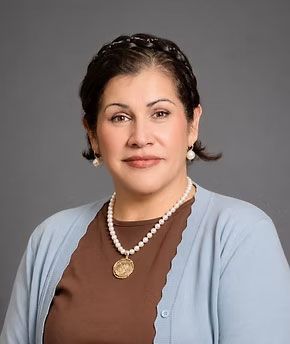 Studio portrait of a woman in a brown top and light blue cardigan, wearing pearl necklace, against a gray background