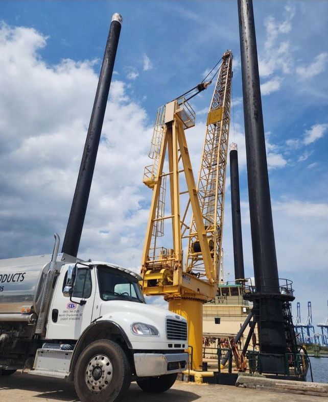 White truck beside a yellow crane and black piles at a construction site under a blue sky
