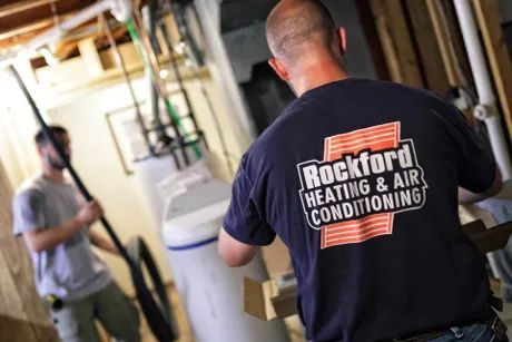 a man wearing a Rockford Heating and Air Conditioning shirt is working on a water heater
