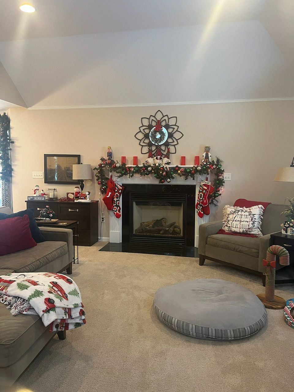 Cozy living room with fireplace, holiday garland, beige chairs, and a gray floor cushion on carpet.