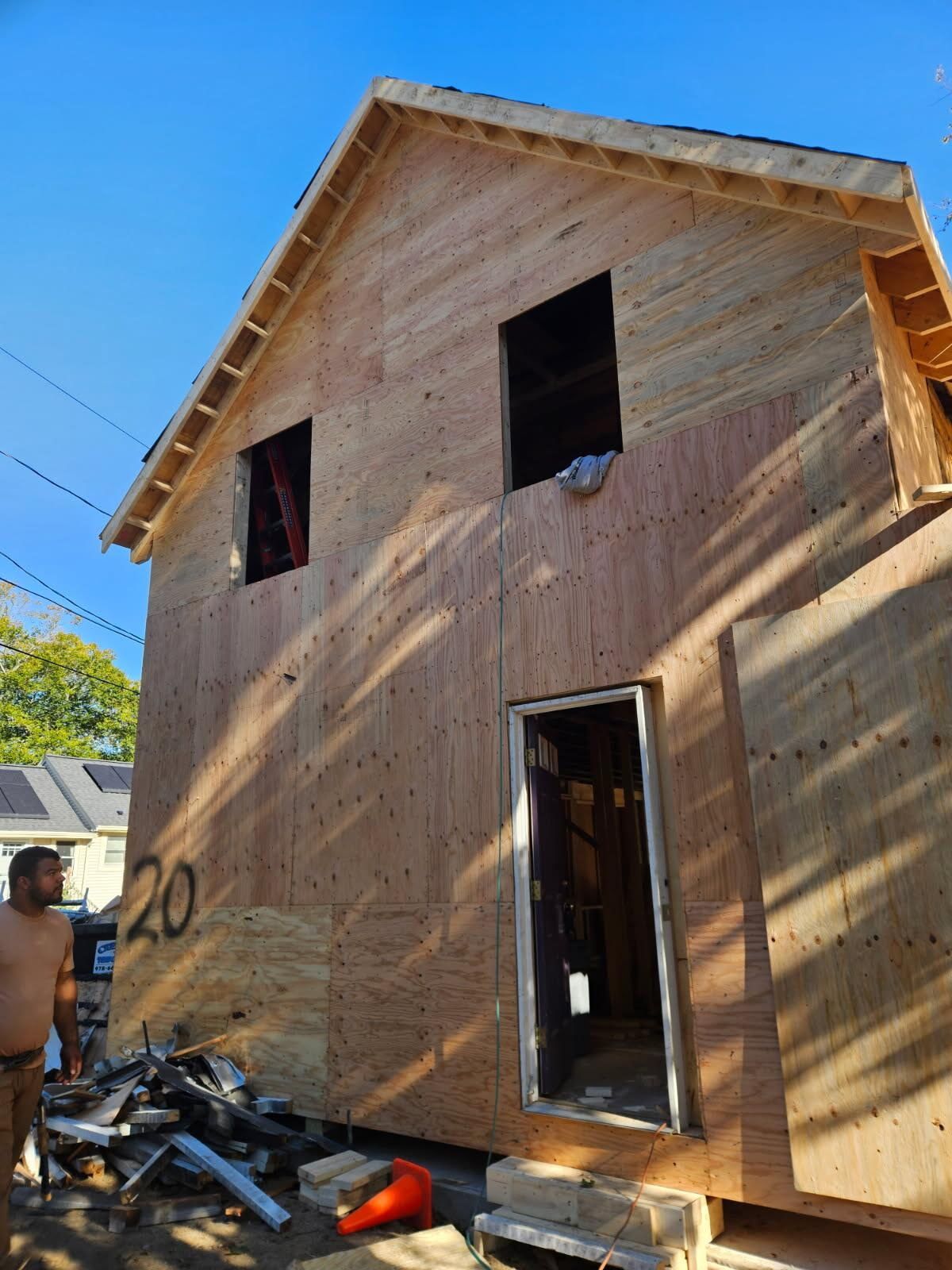 Construction of a two-story building; plywood exterior with door and window openings; man standing nearby.