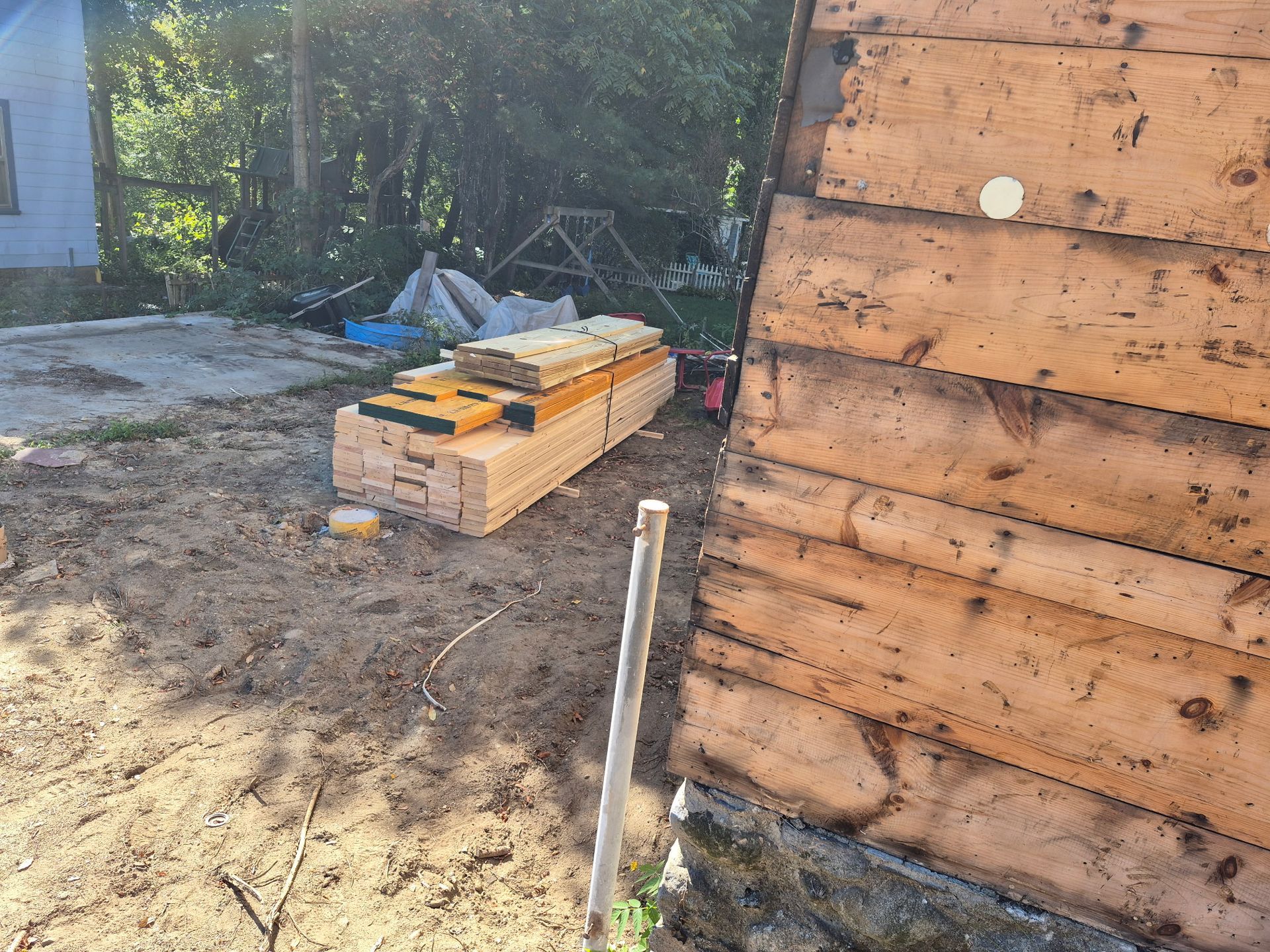 Construction site with stacked lumber, insulation, and a wooden wall. Sand ground, sunny outdoors.