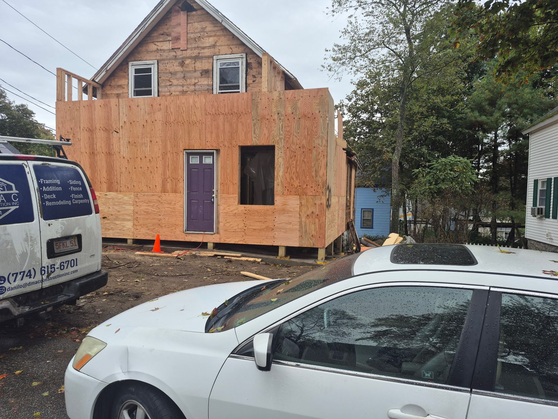 House under construction, unfinished wood exterior, purple door, white cars parked in front, trees.