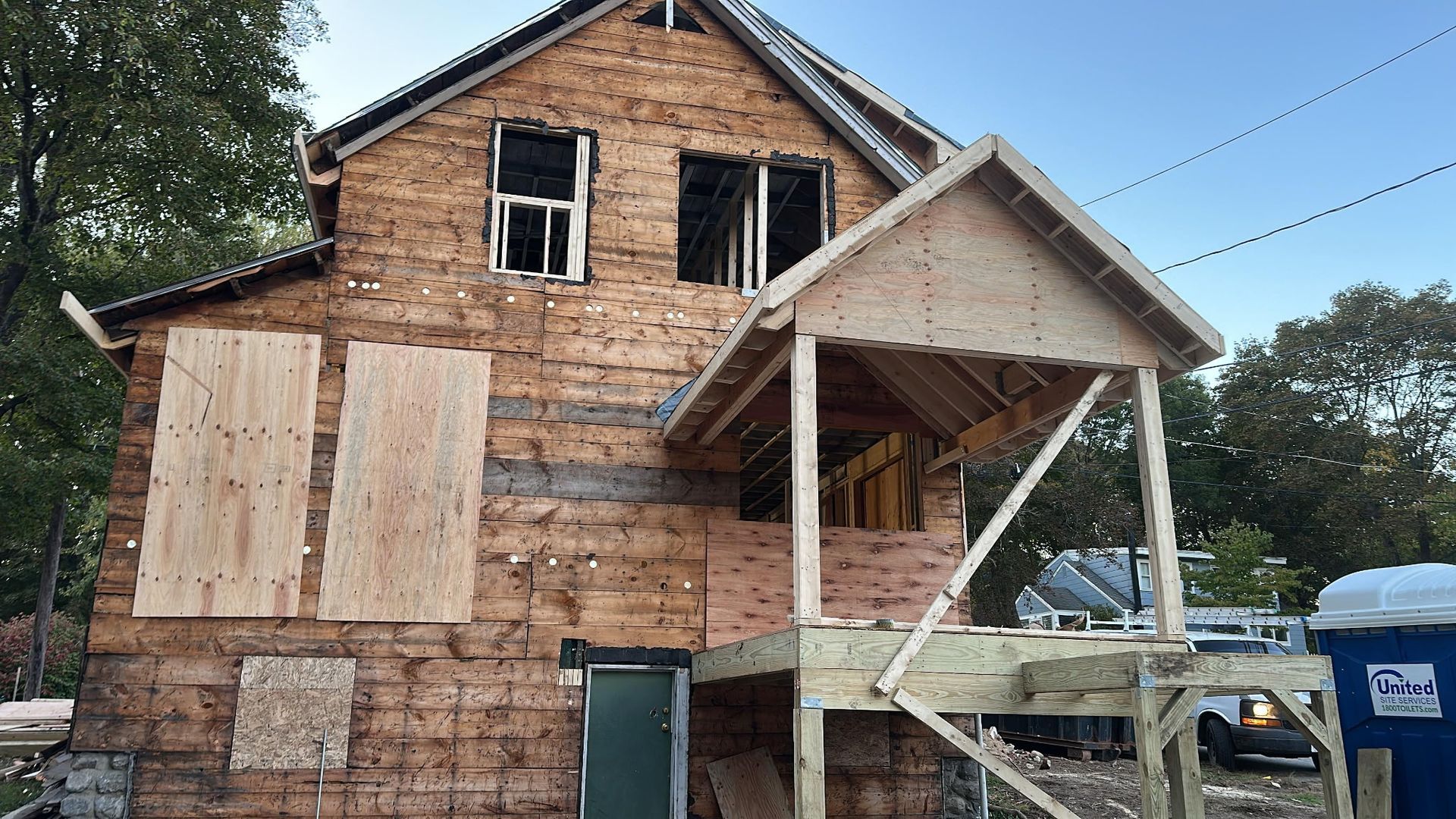 A two-story house under construction with a partially built porch. Wooden panels cover some windows.