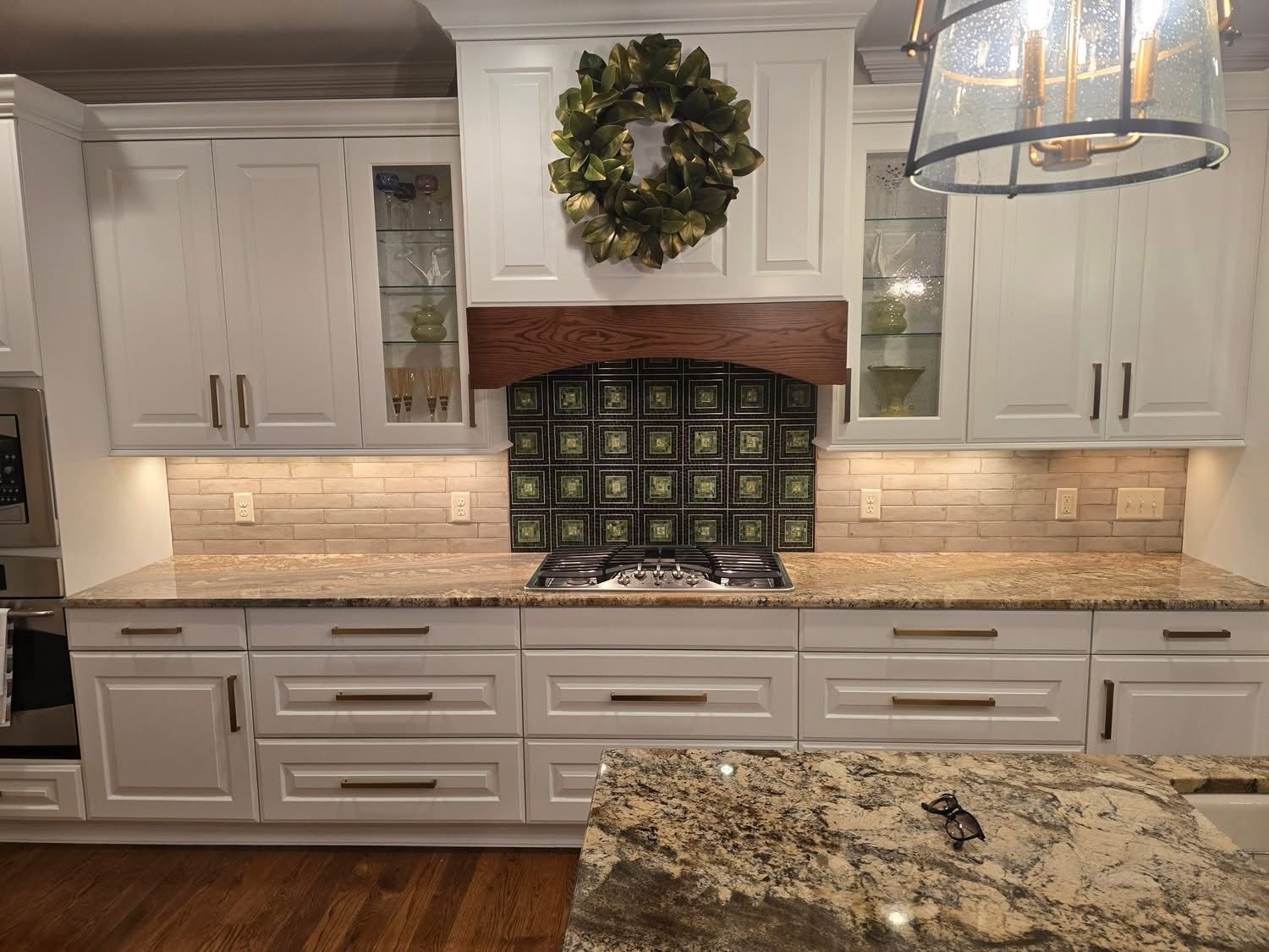 White kitchen cabinets with granite countertops, stovetop, glass block backsplash, and decorative wreath.