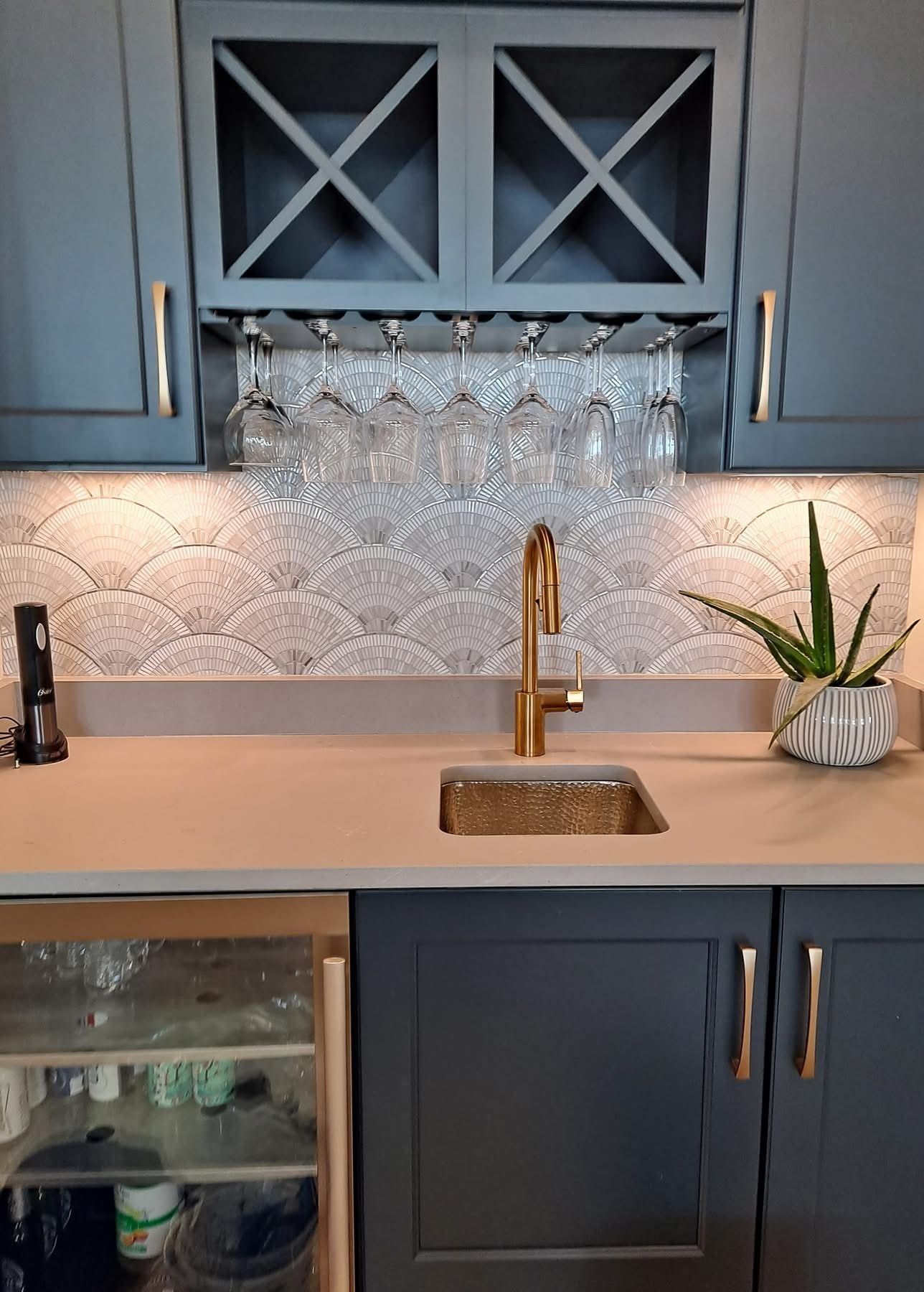 Bar area with a sink, wine glasses, and a decorative tile backsplash. Dark blue cabinets with gold hardware.