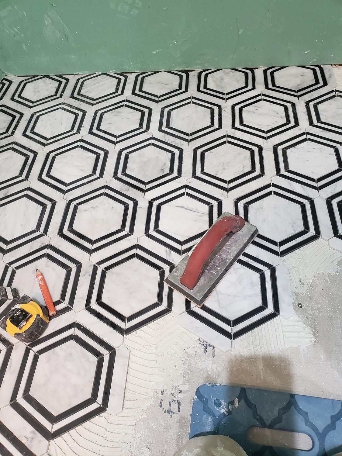 Tiled floor with hexagon pattern in black and white, tools visible: trowel, measuring tape. Green wall in the background.