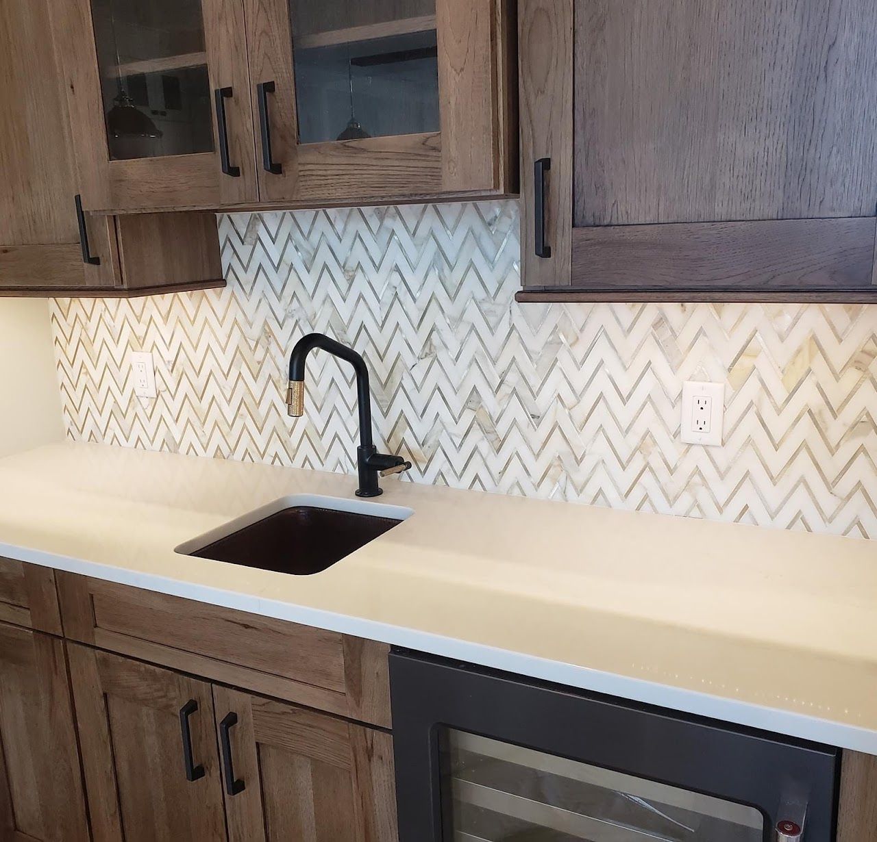 A small kitchen area with a sink, black faucet, and chevron tile backsplash. Wooden cabinets and a wine fridge are visible.
