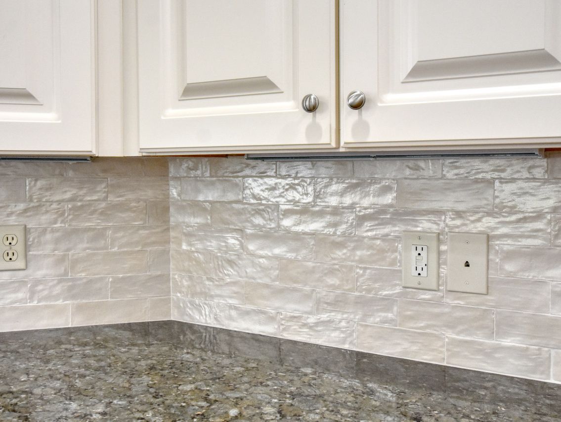 White kitchen cabinets above off-white, textured backsplash and gray countertop. Two electrical outlets present.