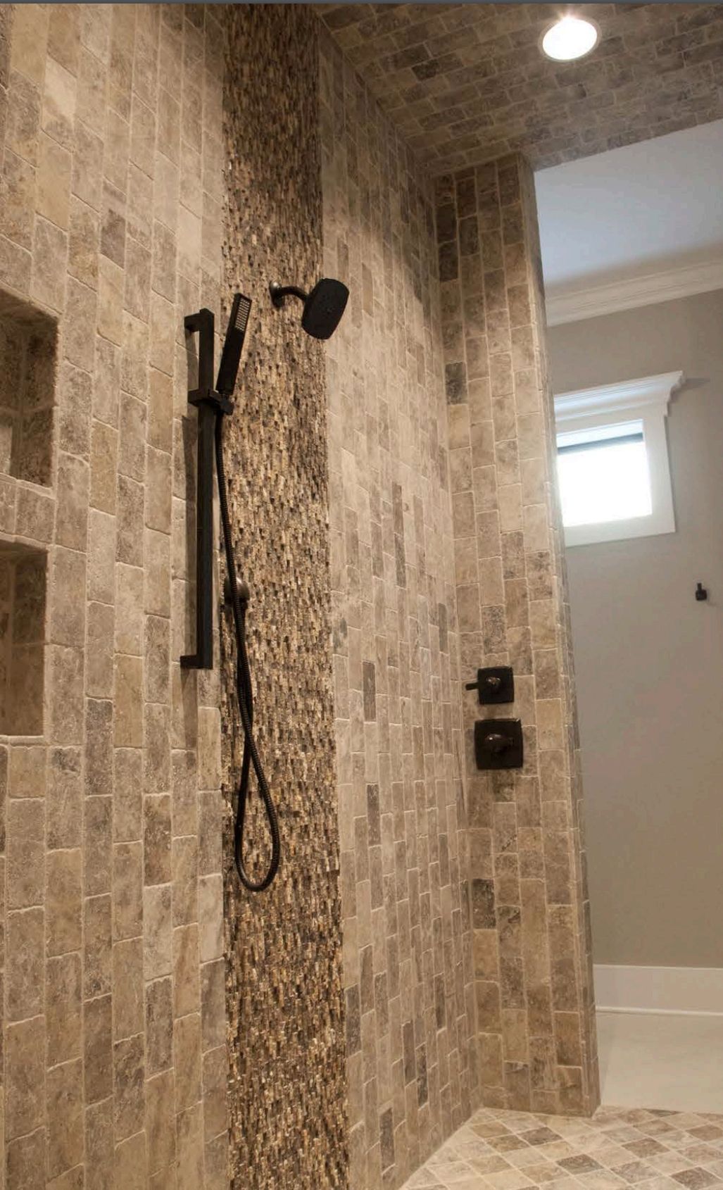 Shower with brown and tan stone tile, a black showerhead, and a small window.