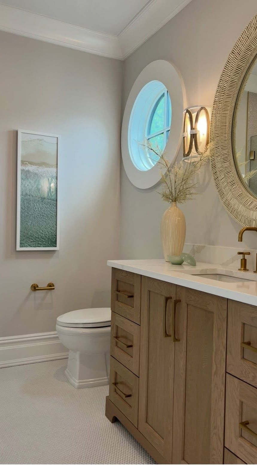 Bathroom with light wood vanity, round window, gold sconce, and oval mirror.