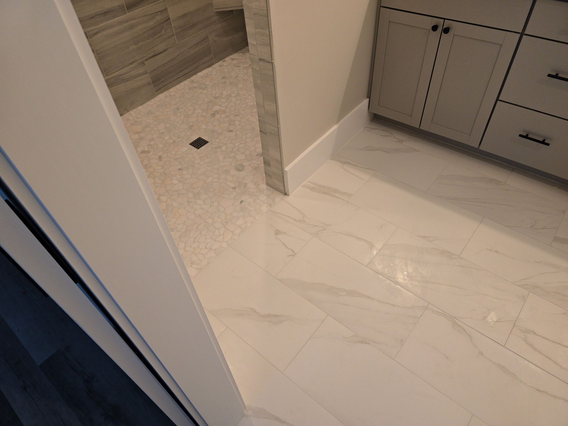 Bathroom with white marble tile floor, shower with pebble floor, and gray cabinet.