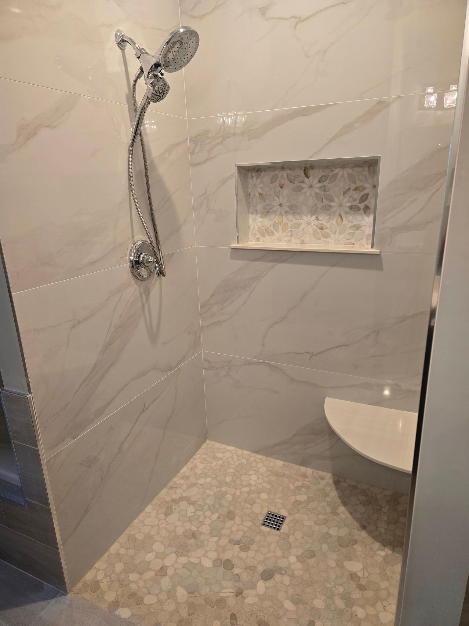 Shower with marble-look tile walls, pebble floor, built-in shelf, and curved bench seat. Chrome shower head and fixtures.