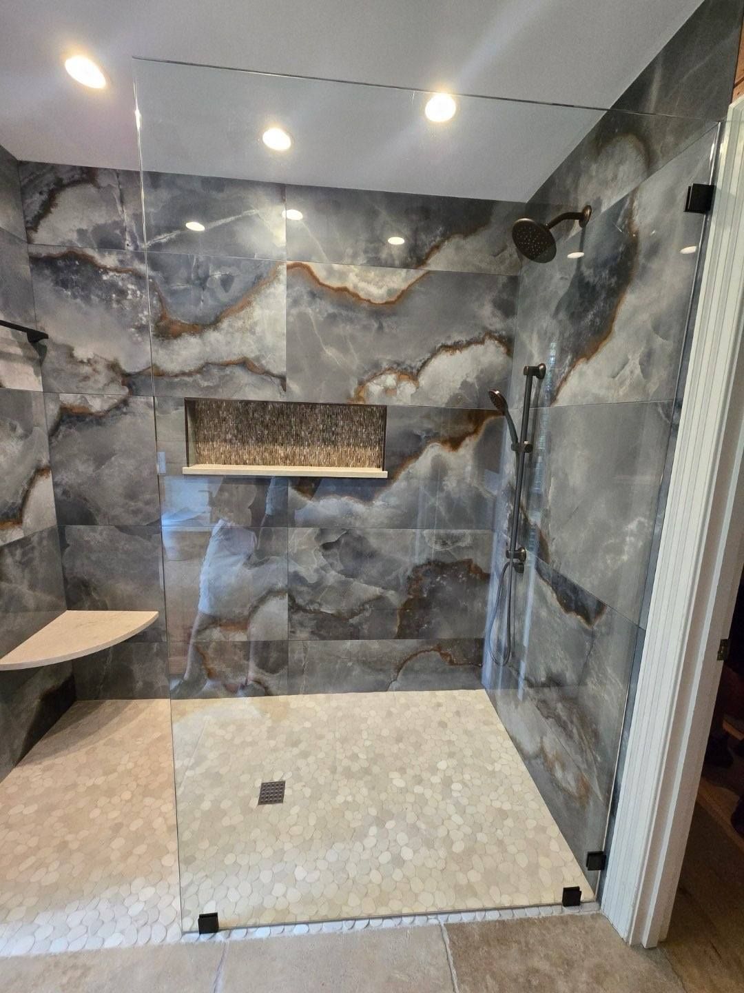 Walk-in shower with grey stone tile walls, glass door, and built-in shelf.